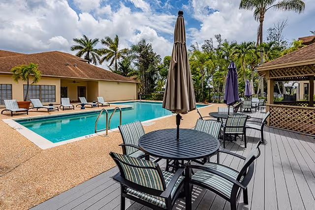 Outdoor swimming pool and patio area with tables, umbrellas, lounge chairs, and a building in the background.
