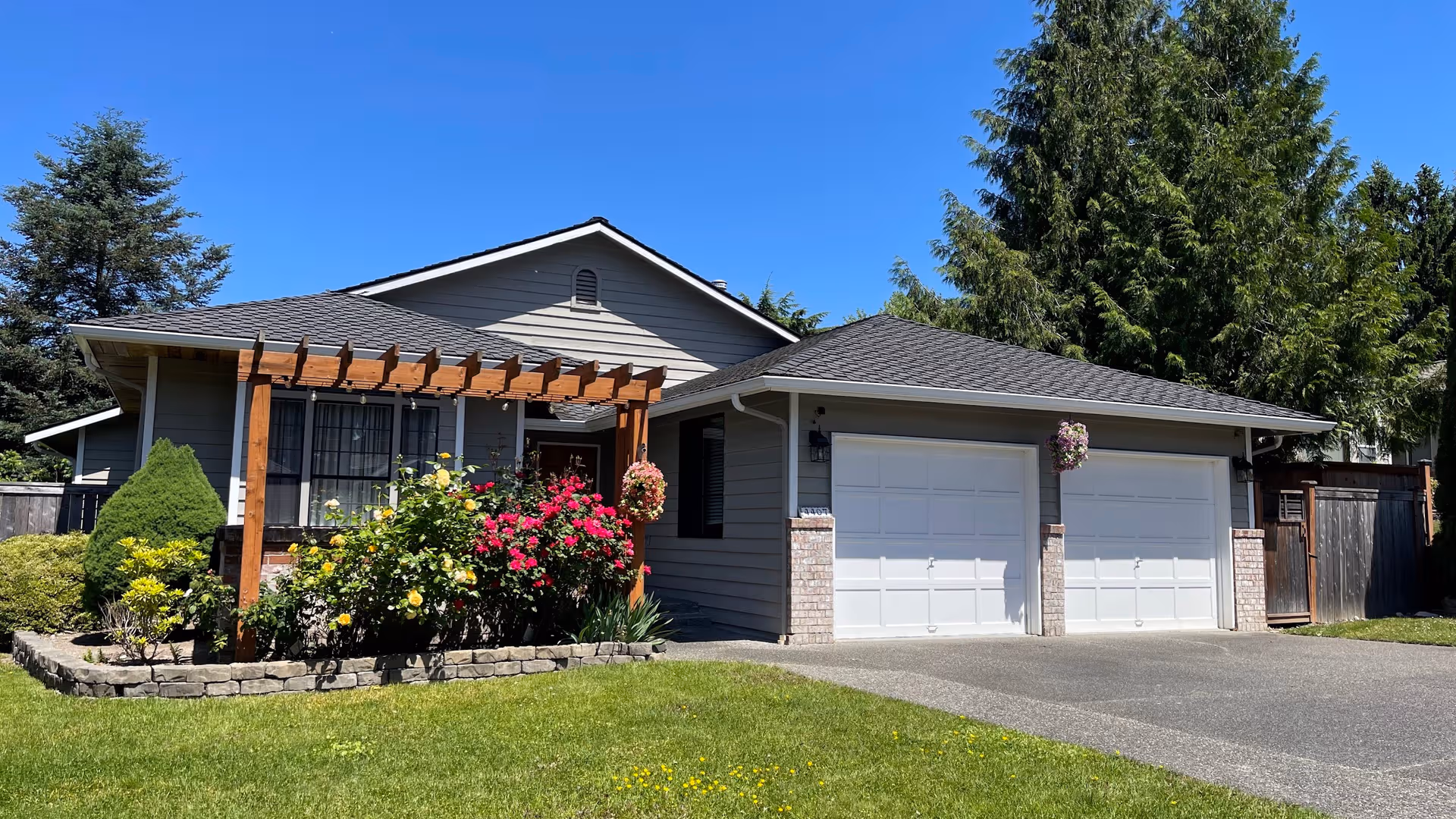 A single-story gray house with a two-car garage, landscaped front yard, and wooden pergola.