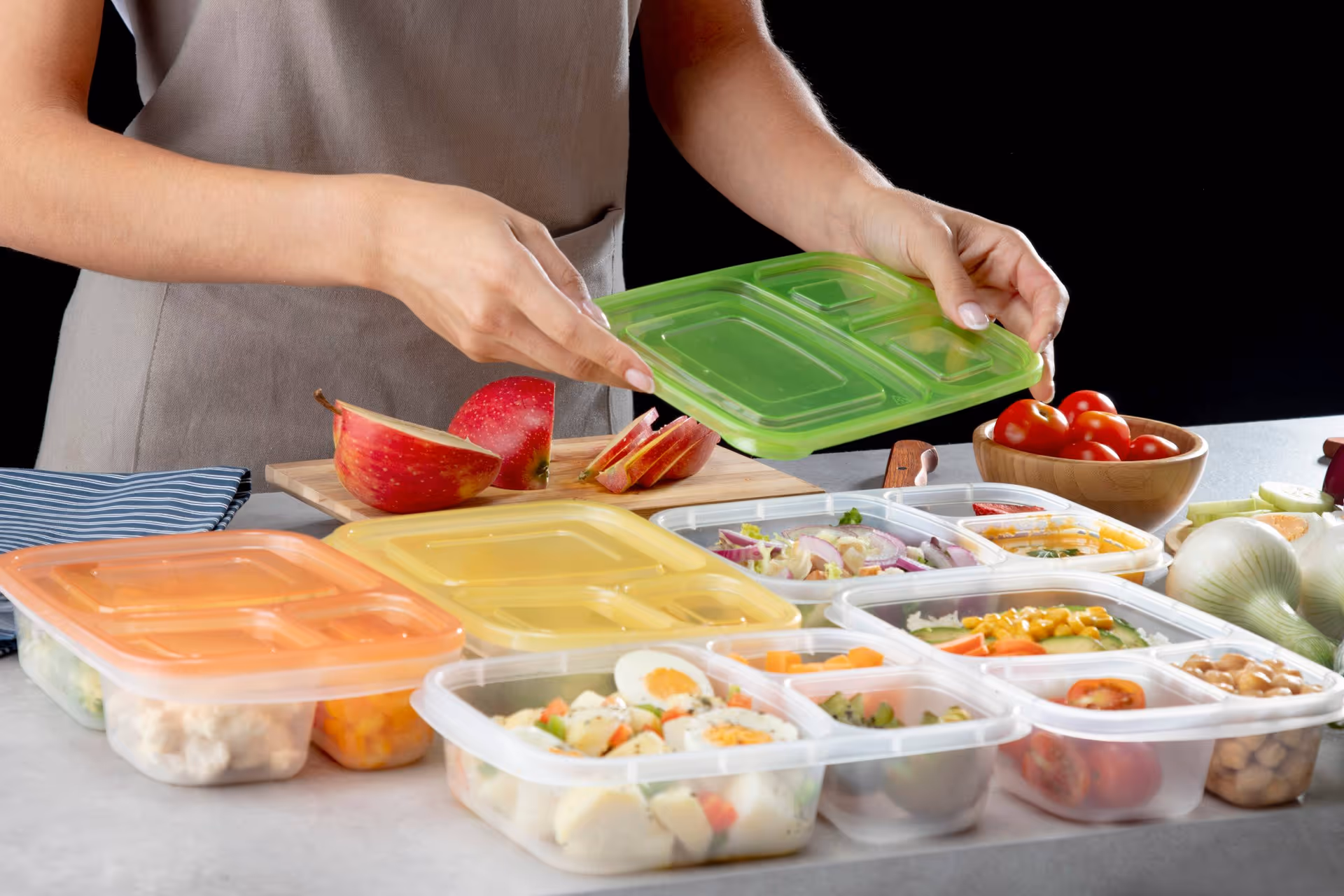 Hands place a green lid on assorted meal-prep containers with produce.