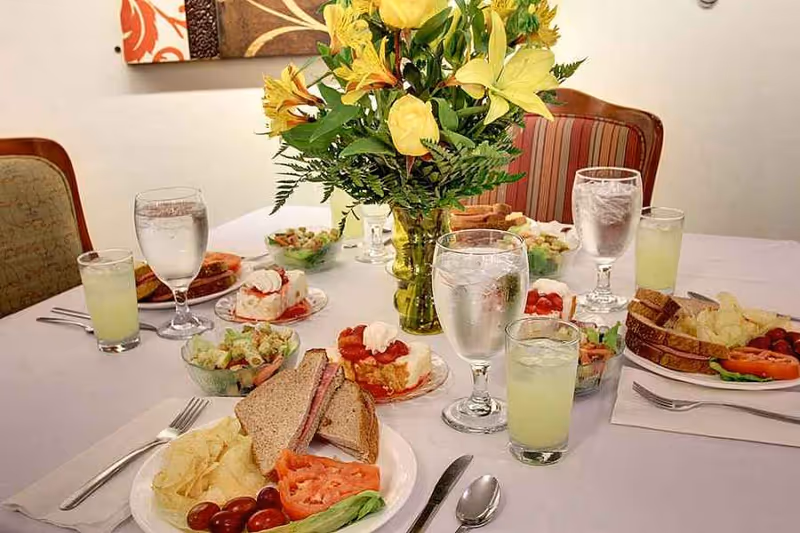 A dining table set with sandwiches, salads, desserts, glasses of water and lemonade, and a vase of yellow flowers.