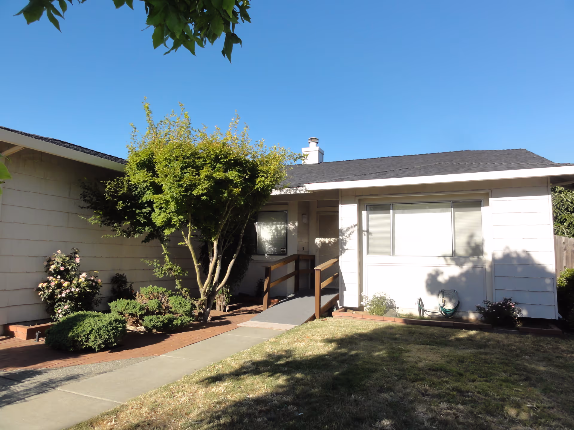 Exterior view of a single-story residential building with white siding, a black roof, and a small ramp leading to the entrance. There are green shrubs and a tree in front of the building, along with a lawn and a clear blue sky.