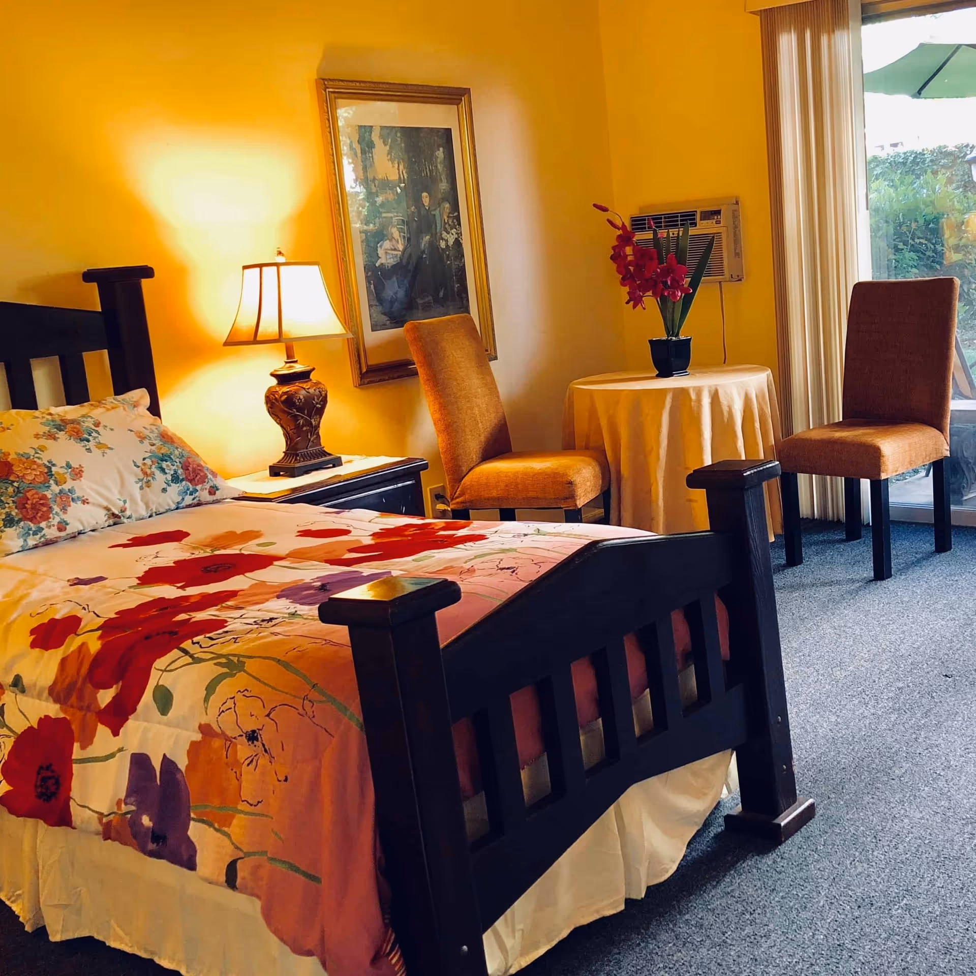 A warmly lit bedroom with a wooden bed dressed in a floral comforter, a bedside lamp and nightstand, framed artwork, and two chairs with a small round table by a sliding glass door.