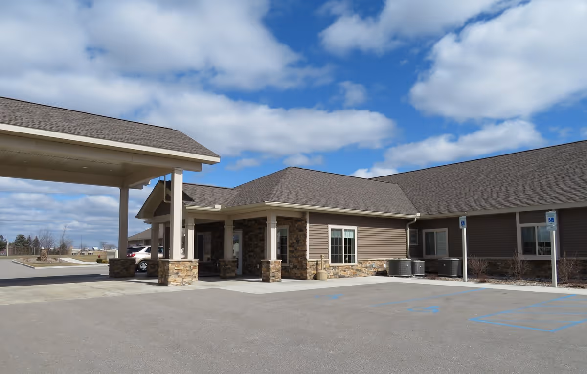 Exterior view of a single-story building with a covered entrance supported by stone pillars, a parking lot with handicap parking spaces, and a partly cloudy blue sky.