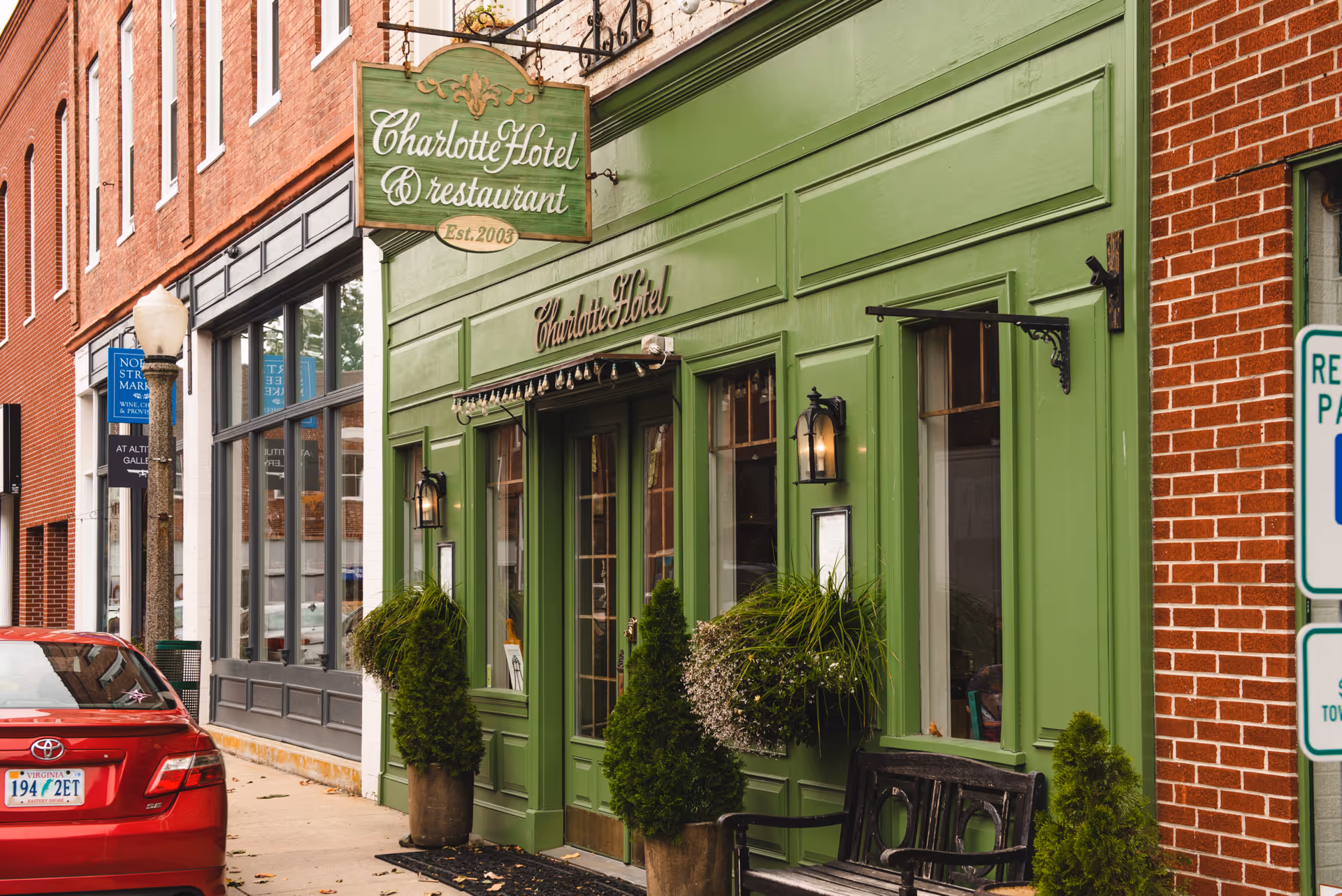 Street view of the Charlotte Hotel & restaurant with a green exterior facade, potted plants, a bench, and a red car parked nearby. The building is located on a sidewalk with other brick buildings and street signs visible.