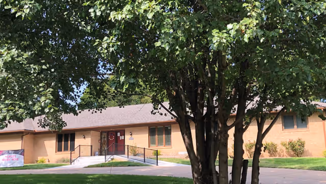 Exterior view of a single-story brick building partially obscured by a large leafy tree in the foreground. The building has a red door with a ramp and steps leading up to it, surrounded by green grass and small shrubs under a clear sky.