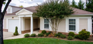 Single-story residential-style building with a covered entry supported by columns, an attached garage, windows, and landscaped lawn with a tree.