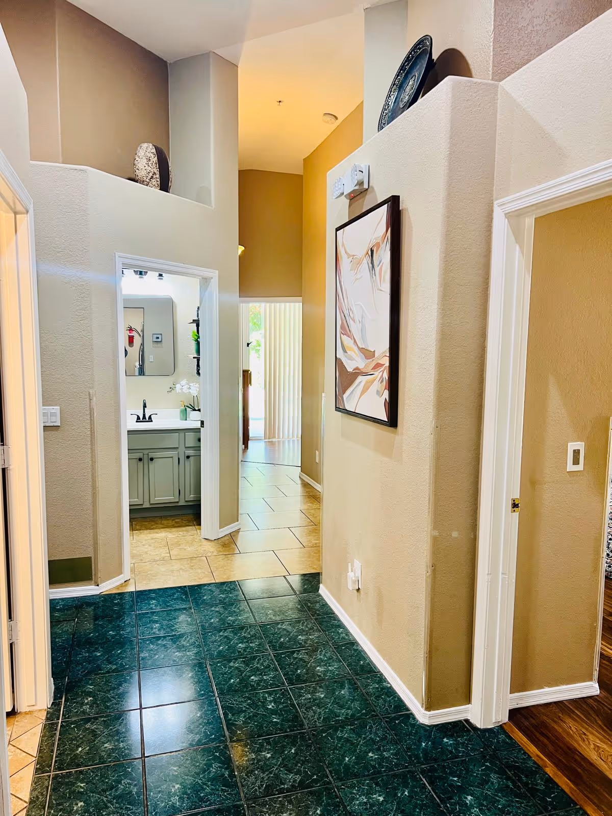 Interior hallway of a senior living facility with green tiled flooring leading to a bathroom on the left and other rooms on the right. The walls are beige with a framed abstract painting hanging on the right wall. The bathroom has a gray vanity with a mirror and a black faucet. The hallway opens up to a brighter area with beige tiled flooring and vertical blinds covering a window or door.