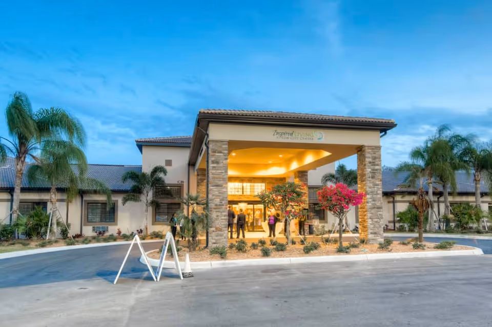 Front entrance of a senior living facility with a covered porte-cochère, palm trees, and people near the lit doorway.