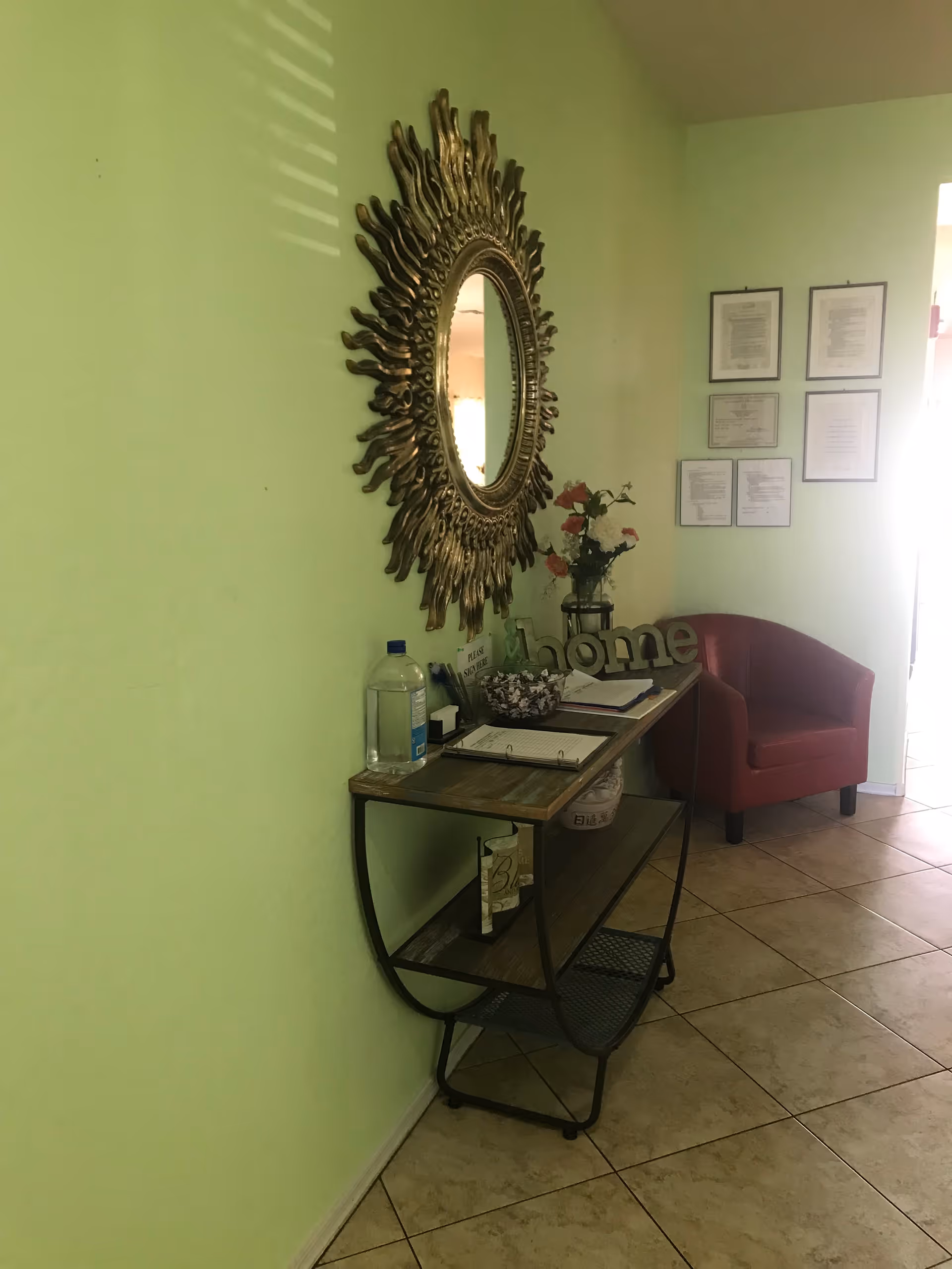 A small entryway or waiting area with a decorative sunburst mirror on a light green wall, a narrow wooden console table with a water bottle, a flower vase, and a 'home' decoration on top. There is a red armchair in the corner and framed documents or pictures on the adjacent wall.