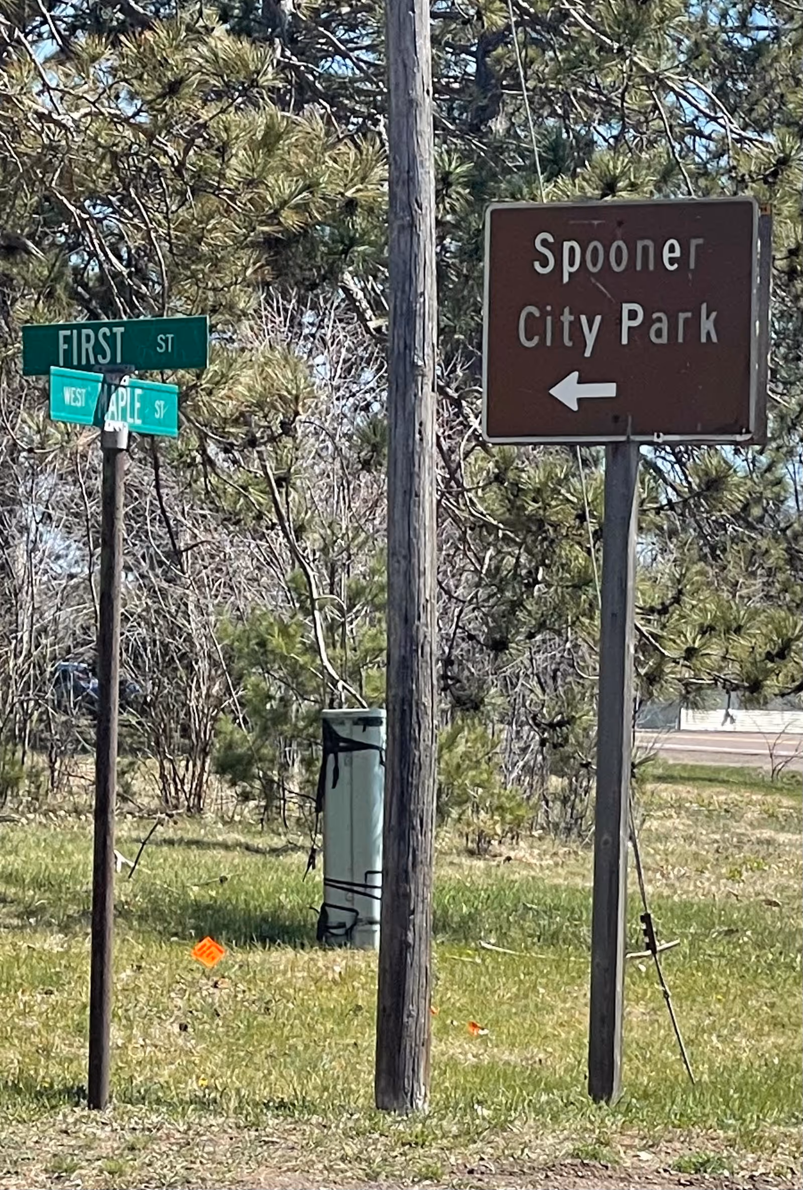 Street signs at an intersection with a wooden utility pole in the foreground. One green sign indicates the intersection of First Street and Maple Street, while a brown sign points left towards Spooner City Park. Trees and grass are visible in the background.