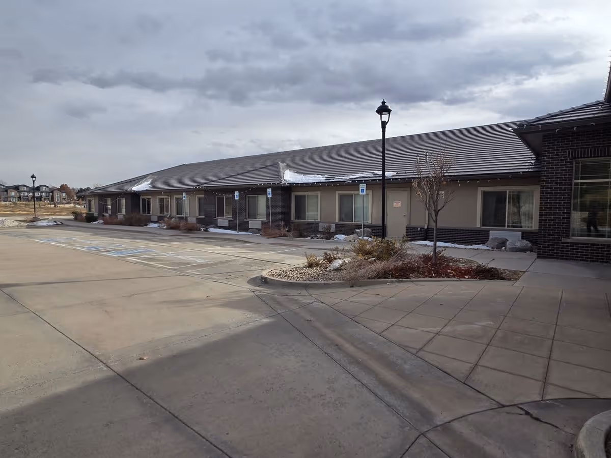 A single-story memory care building with a row of windows, adjacent parking with handicap spaces, lamp posts, and a cloudy sky.