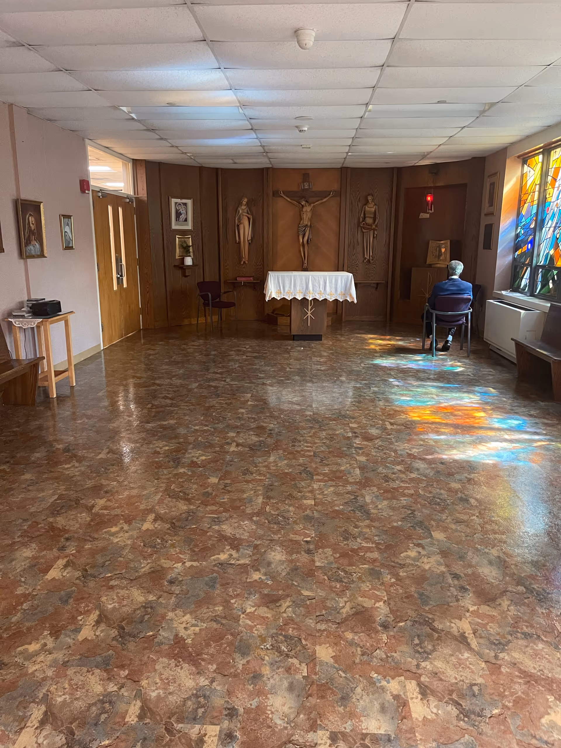 A chapel interior with altar, crucifix, stained glass, and a seated resident.