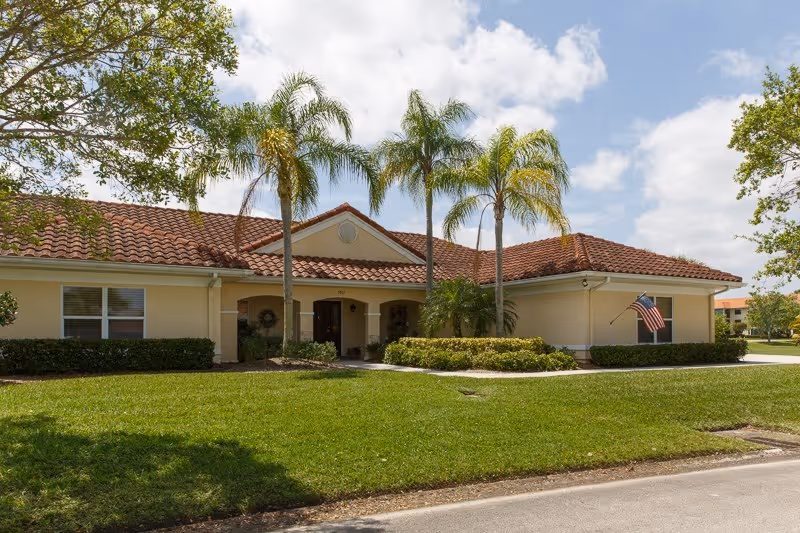 Single-story cream-colored building with a red tiled roof, palm trees, manicured lawn, and an American flag at the entrance.
