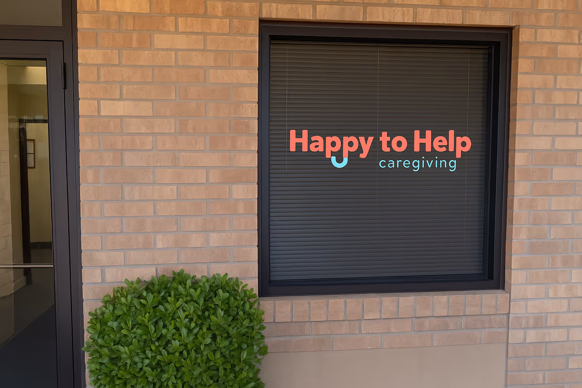 Brick exterior with a window sign reading 'Happy to Help caregiving' and a shrub.