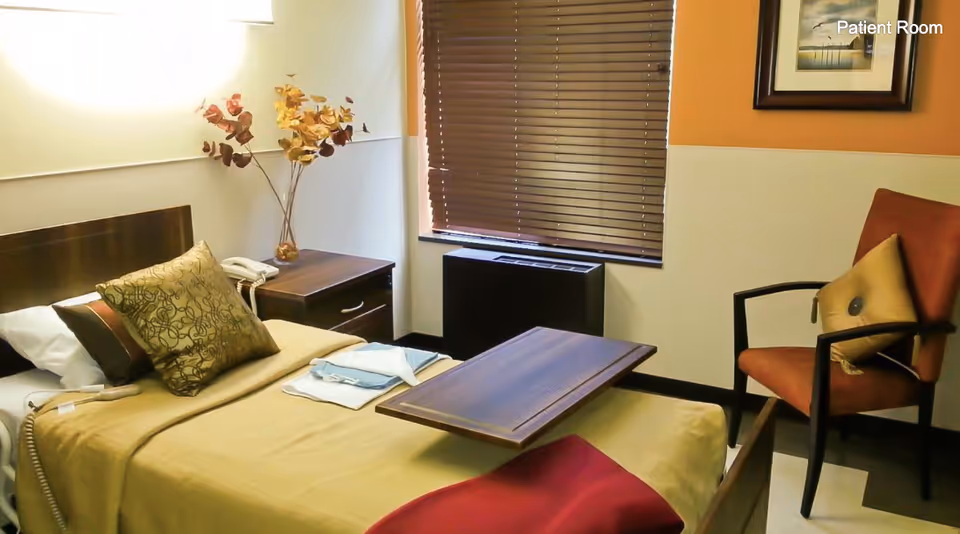 A patient room with a single bed covered in beige and brown bedding, a decorative pillow, and a wooden overbed table. There is a nightstand with a telephone and a vase with artificial flowers next to the bed. A window with closed brown blinds and a heating/cooling unit beneath it is visible. An orange armchair with a cushion is placed near the bed, and a framed picture hangs on the wall.