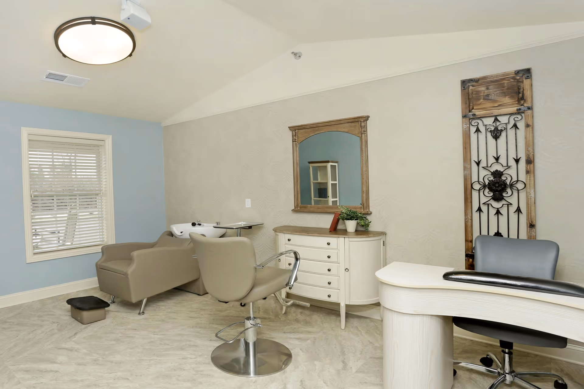 A well-lit salon area in an assisted living facility featuring a beige salon chair with a footrest, a hair washing station with a reclining chair, a white dresser with a wooden framed mirror above it, a small plant and a book on the dresser, a window with blinds, and a decorative wooden panel with wrought iron design on the wall.