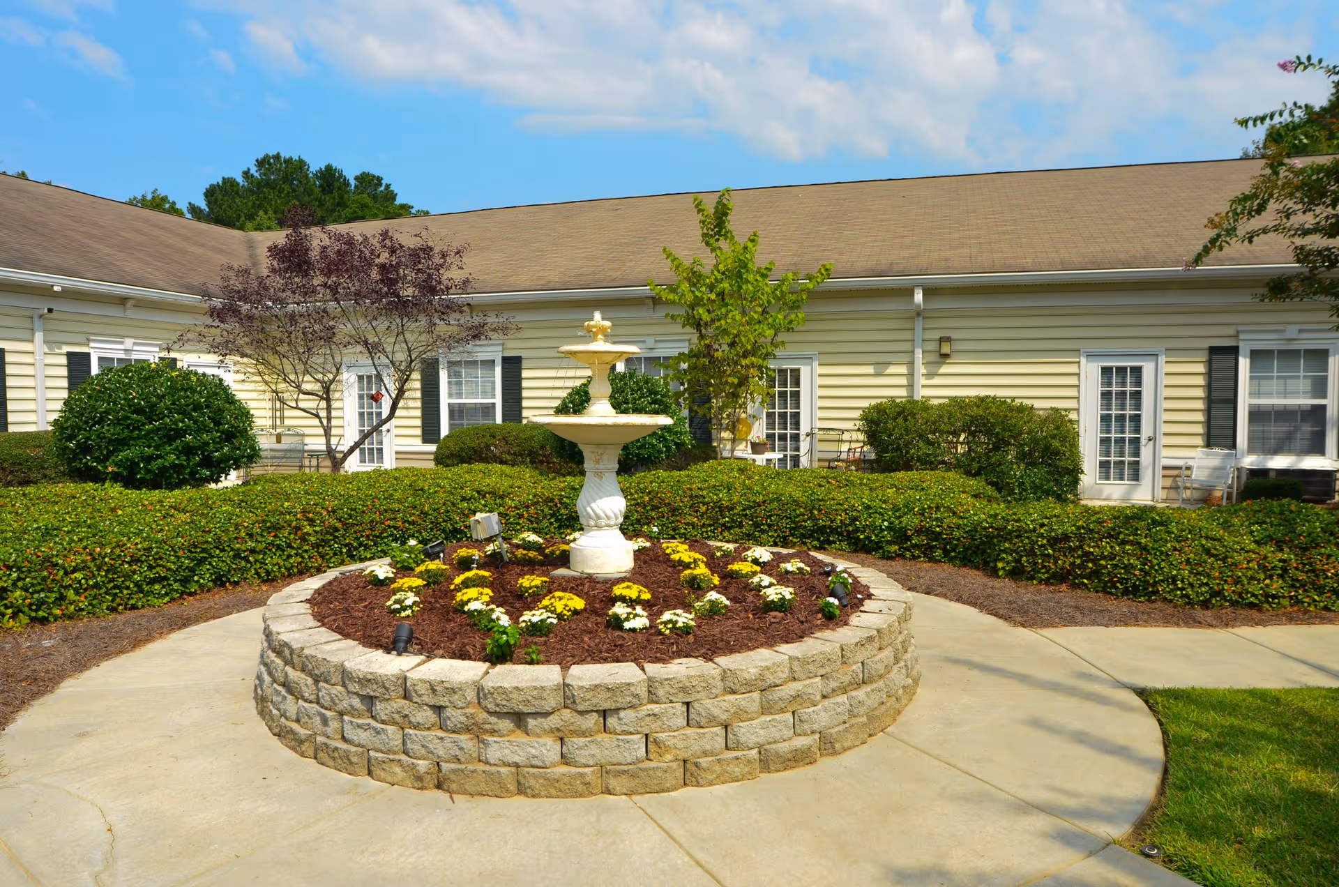 Front exterior of a beige senior living building with a circular stone fountain.
