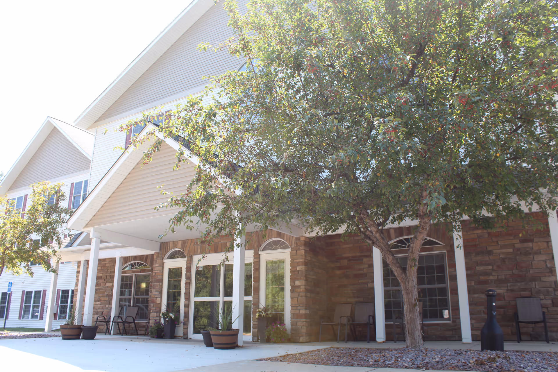 Exterior view of a senior living facility entrance with a covered porch supported by white columns, stone and siding facade, several potted plants, outdoor chairs, and a tree with green leaves in front.