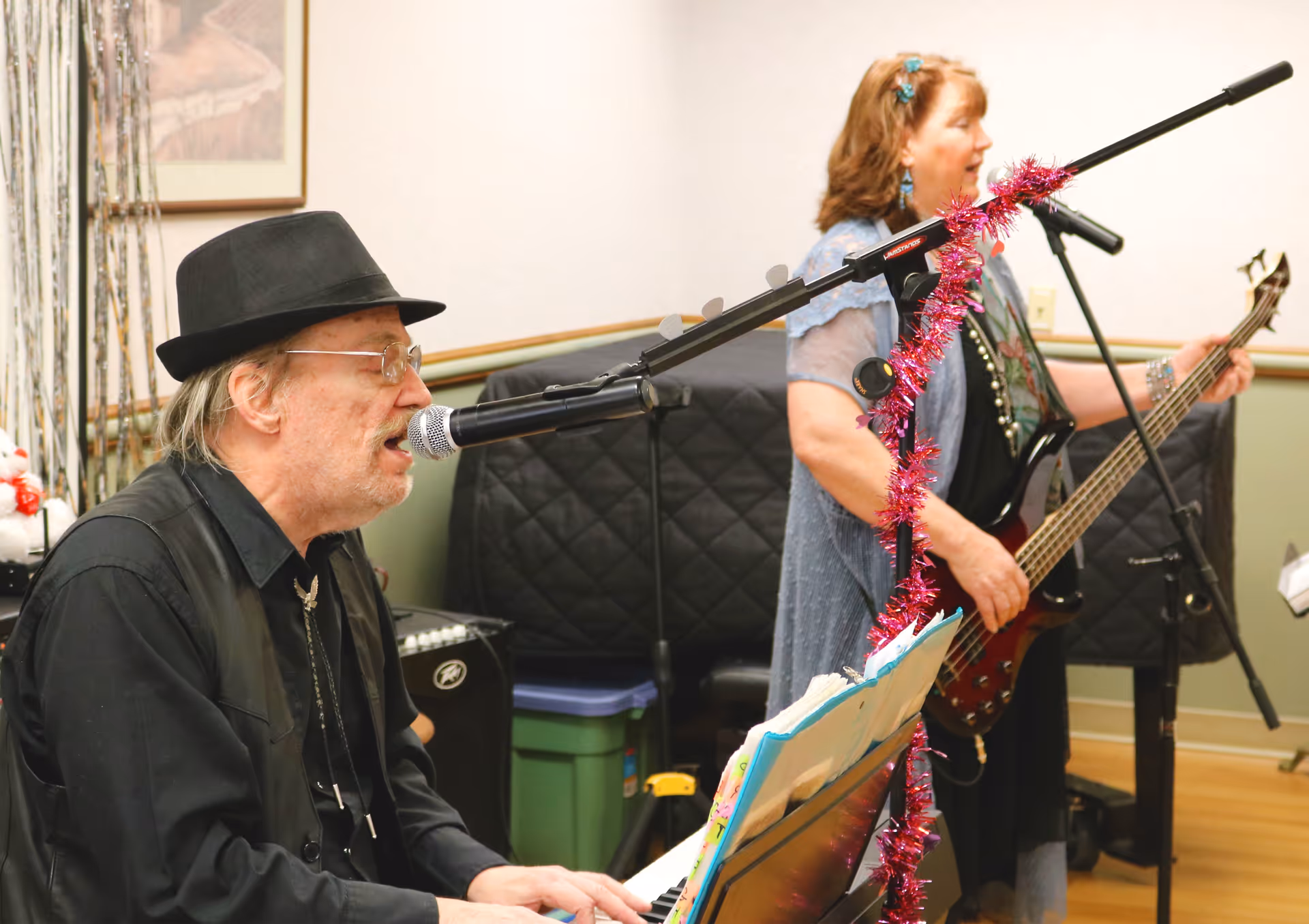 An elderly man wearing a black hat and glasses is playing the keyboard and singing into a microphone. A woman standing next to him is playing a bass guitar and singing into another microphone. The microphone stand is decorated with pink tinsel. They are performing indoors in a room with light-colored walls and wooden flooring.