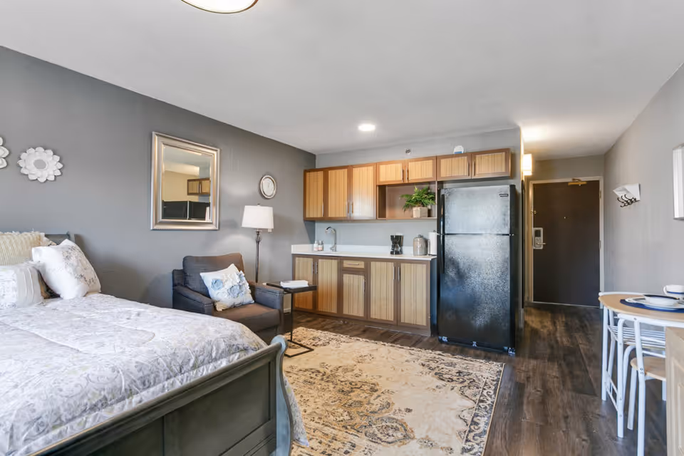 Interior view of a senior living facility room featuring a bed with pillows and a patterned bedspread, a dark armchair with a decorative pillow, a floor lamp, a wall mirror, a clock, a kitchenette with wooden cabinets, a black refrigerator, a coffee maker, and a small dining table with two place settings. The room has wood flooring and a patterned area rug.