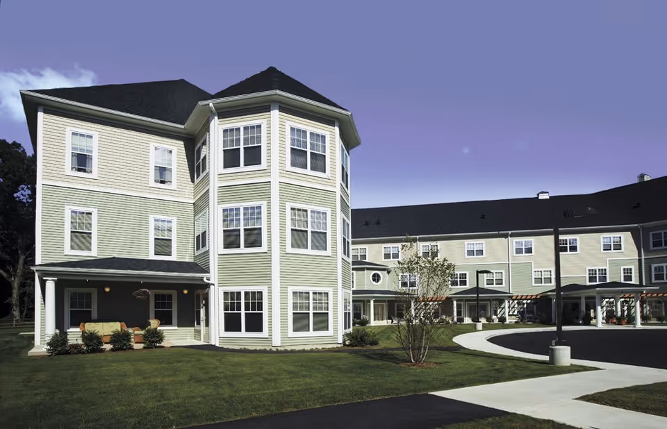 Exterior view of a large, multi-story assisted living facility building with light green and beige siding, multiple windows, and a covered porch area with outdoor seating. The building is surrounded by a well-maintained lawn and paved walkways under a clear sky.