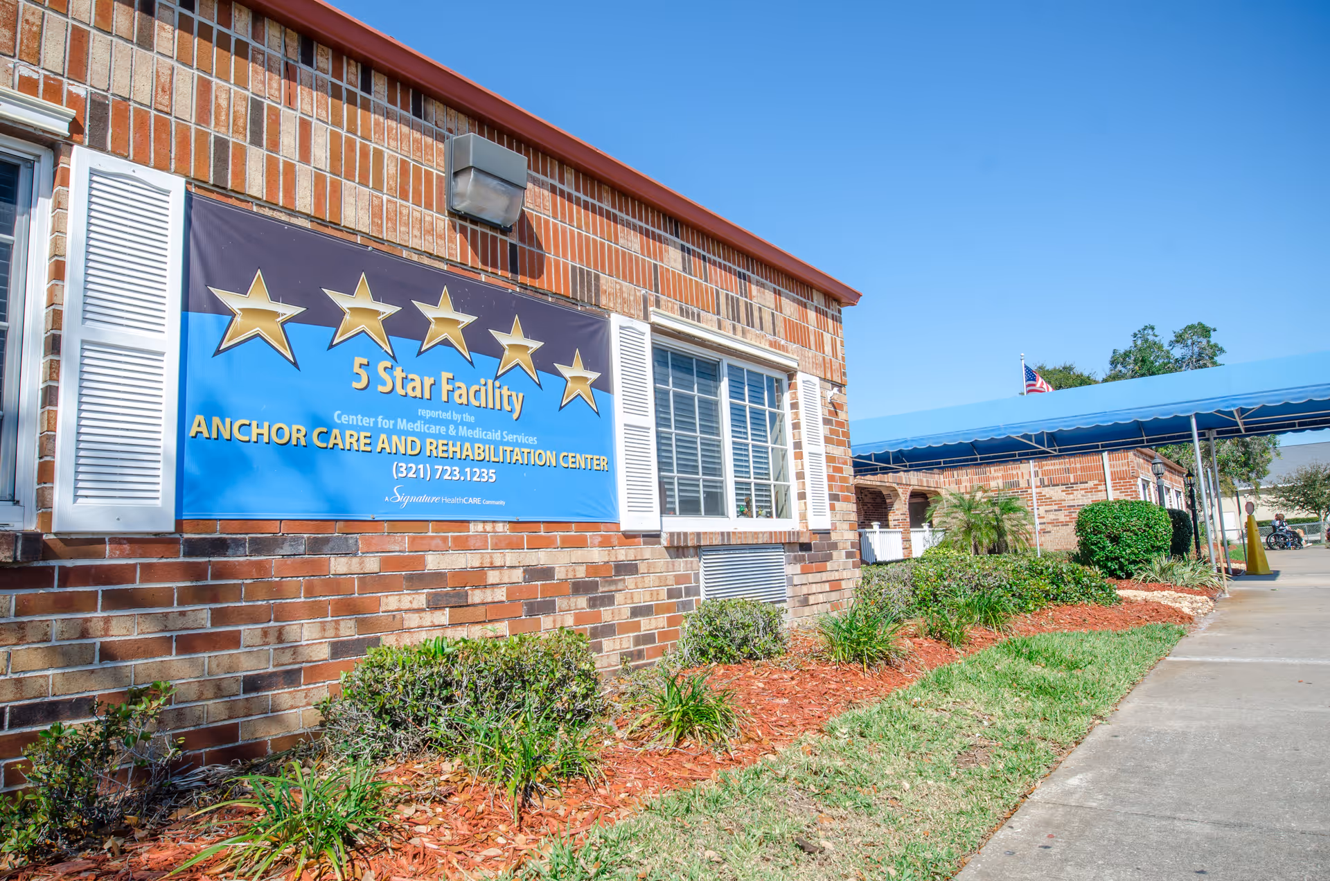 Exterior view of a brick building with a large blue sign displaying five gold stars and the text '5 Star Facility reported by the Center for Medicare & Medicaid Services ANCHOR CARE AND REHABILITATION CENTER (321) 723.1235'. The building has white window shutters and a blue awning covering a walkway. There are bushes and landscaping with mulch along the building.