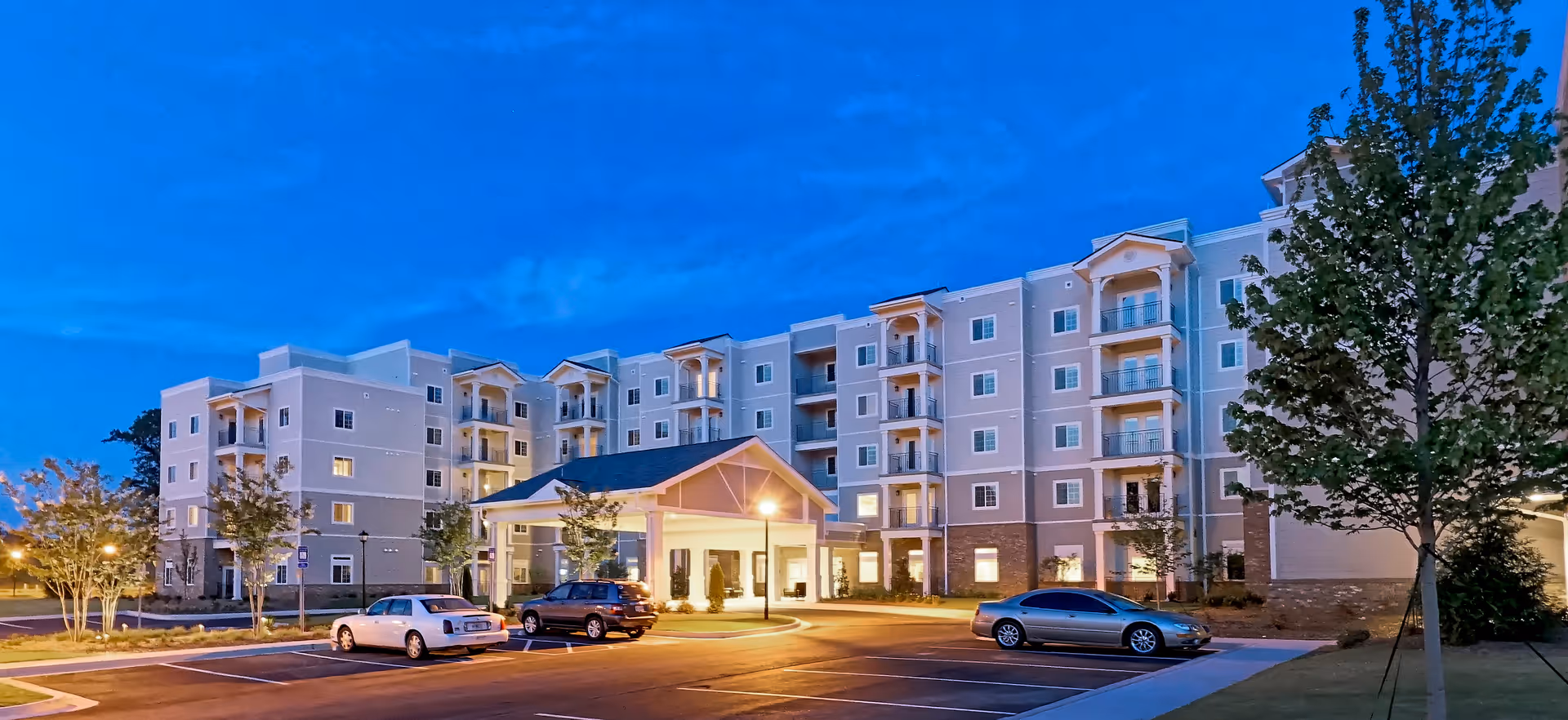 Senior living building with a lit entrance canopy and parked cars at dusk.