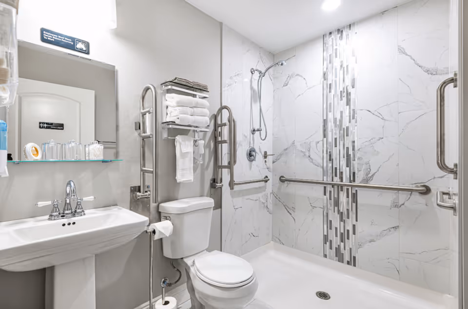 A clean and modern bathroom with a white pedestal sink, a toilet, and a walk-in shower with marble-patterned tiles and a vertical strip of mosaic tiles. The shower has a handheld showerhead and multiple grab bars for accessibility. Above the toilet, there is a metal rack holding neatly folded white towels. A mirror and a glass shelf with toiletries are mounted on the wall above the sink.