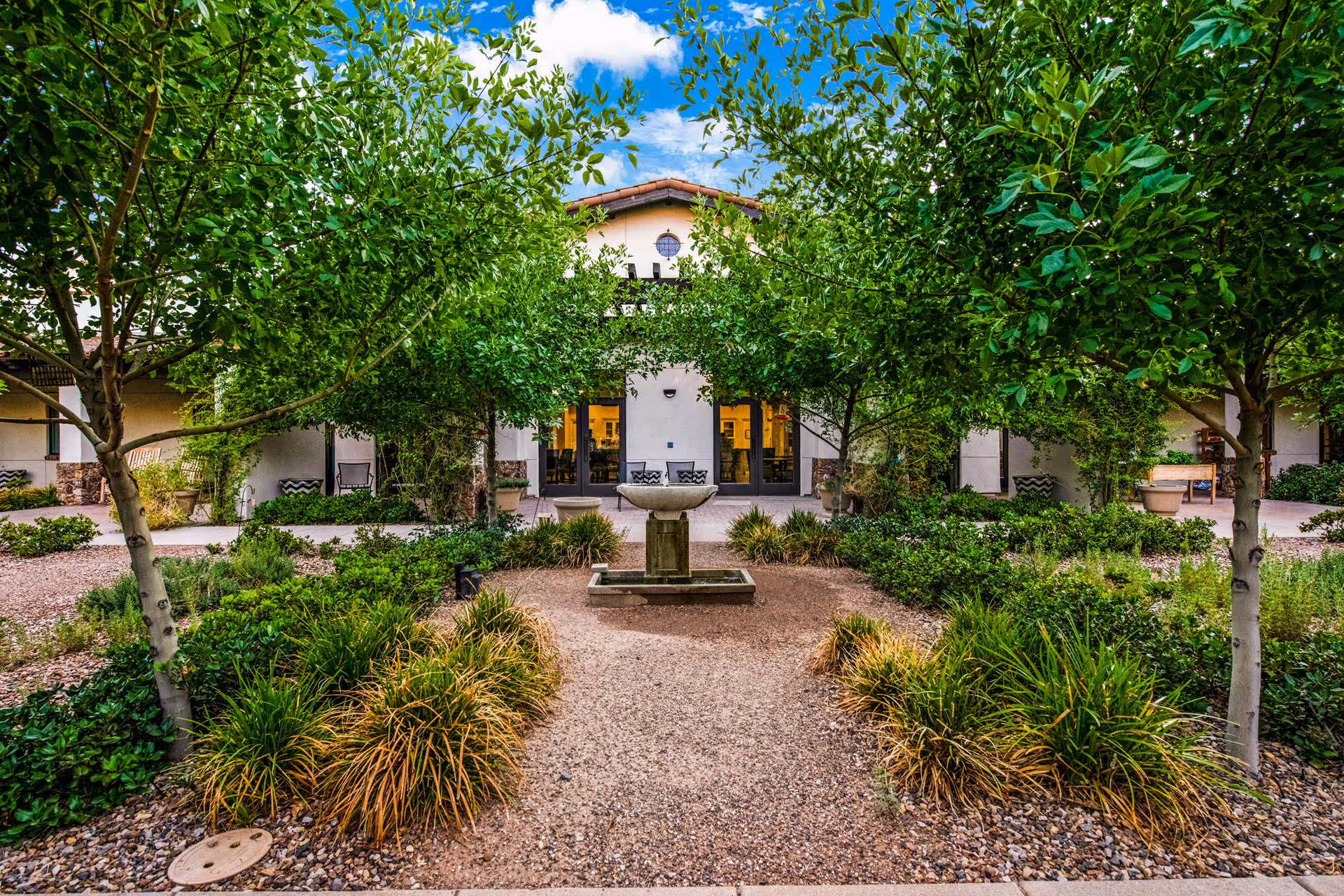 A tree-lined courtyard with a central stone fountain leading to the entrance of a Mediterranean-style building.