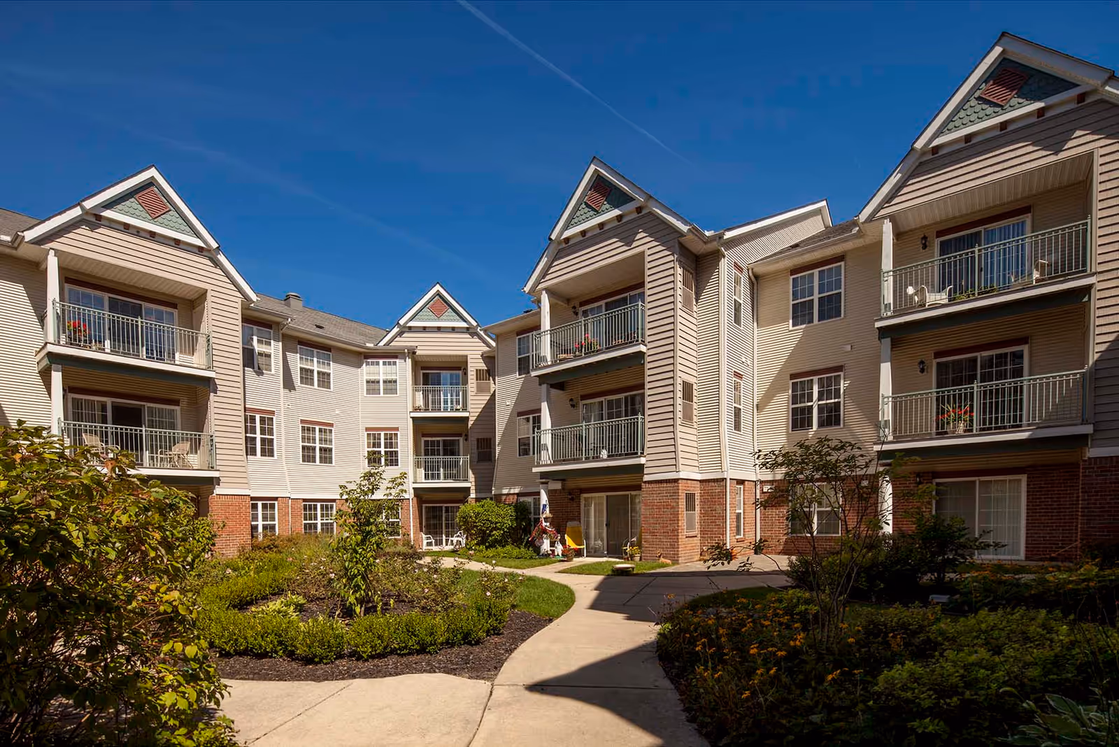 Exterior view of a multi-story residential building with balconies overlooking a landscaped courtyard with pathways, shrubs, and small trees under a clear blue sky.