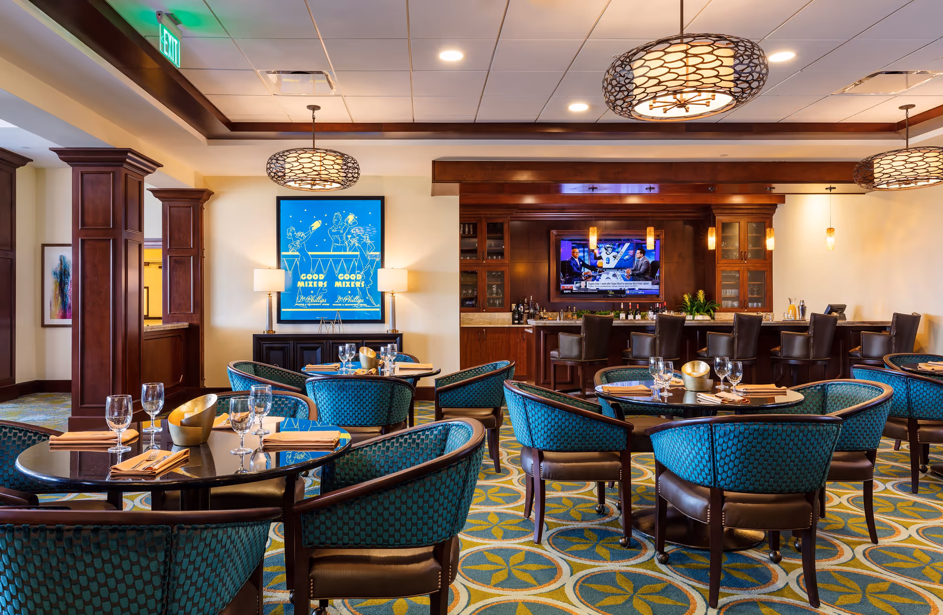 A stylish dining area with round tables set with glasses and napkins, surrounded by teal upholstered chairs. The room features a bar with high chairs, a mounted TV, warm wooden accents, and modern pendant lighting. A colorful carpet with geometric patterns covers the floor, and a bright blue framed poster is displayed on the wall.