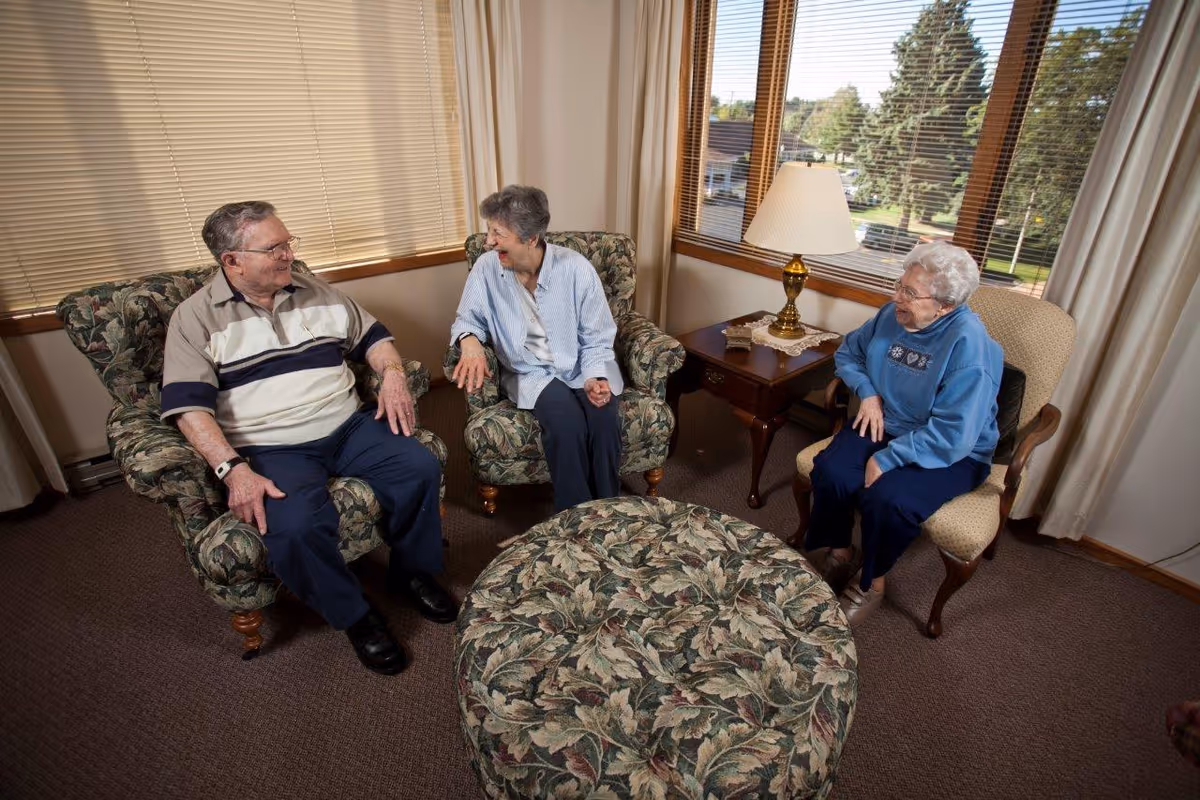 Three elderly people sitting and chatting in a cozy living room with floral patterned armchairs and ottoman, a wooden side table with a lamp, and large windows with blinds and curtains.