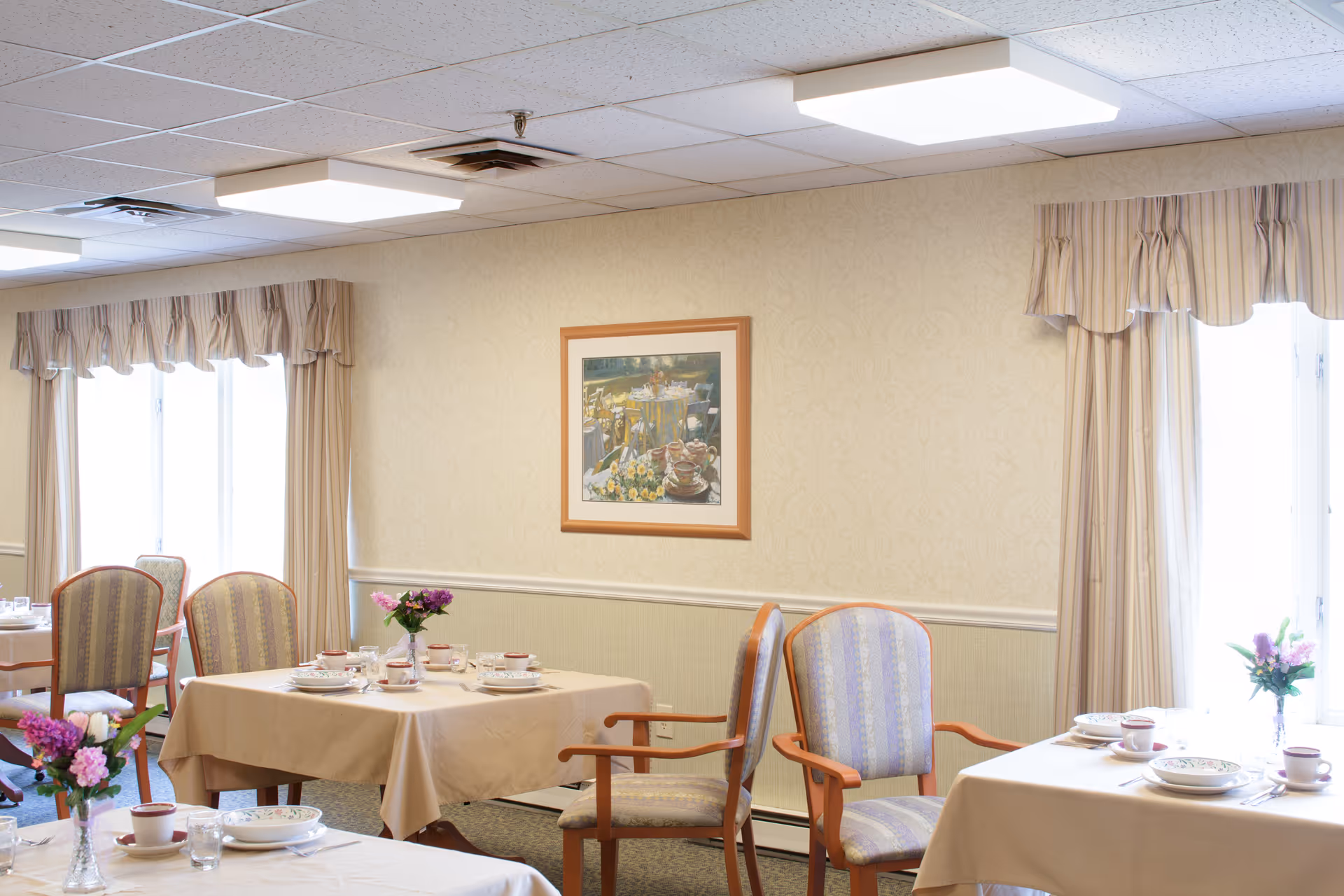 Bright dining room with tables set for meals, upholstered chairs, floral centerpieces, framed artwork, and large curtained windows.