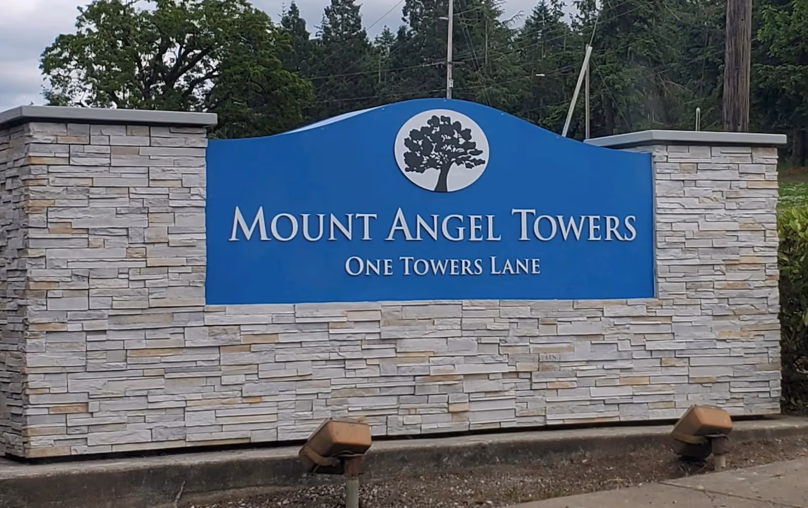 Stone and blue entrance sign that reads "Mount Angel Towers One Towers Lane" with trees in the background.