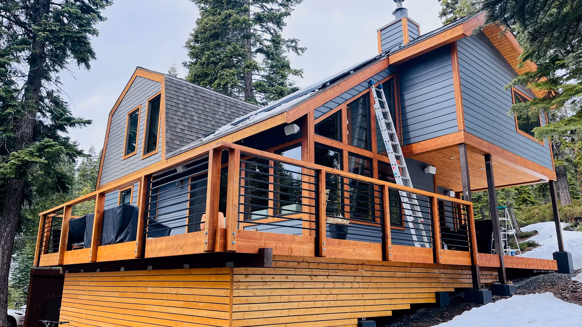 Blue modern house exterior with a wooden deck, railing, and ladder.