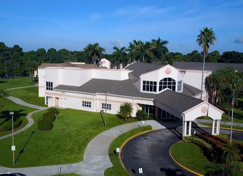 A large light-colored senior living facility exterior with curved driveway and palm trees.