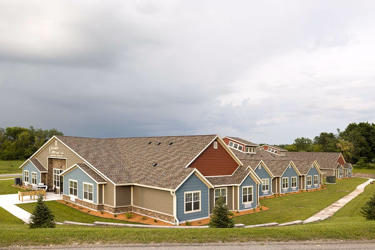 Exterior view of Creamery Creek Senior Living Viroqua - Stoddard building, a single-story structure with multiple gabled roofs, beige and blue siding, and a stone accent near the entrance. The building is surrounded by green grass, small trees, and a paved walkway under a cloudy sky.