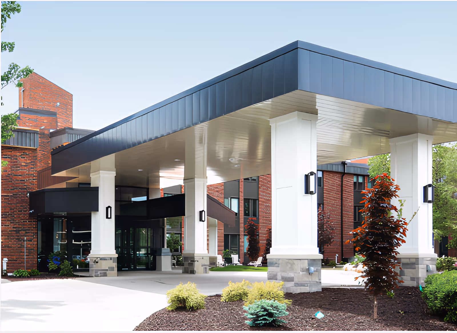 Entrance area of a senior living facility with a covered drop-off zone supported by white columns. The building features red brick walls and large windows, surrounded by landscaped bushes and small trees under a clear sky.