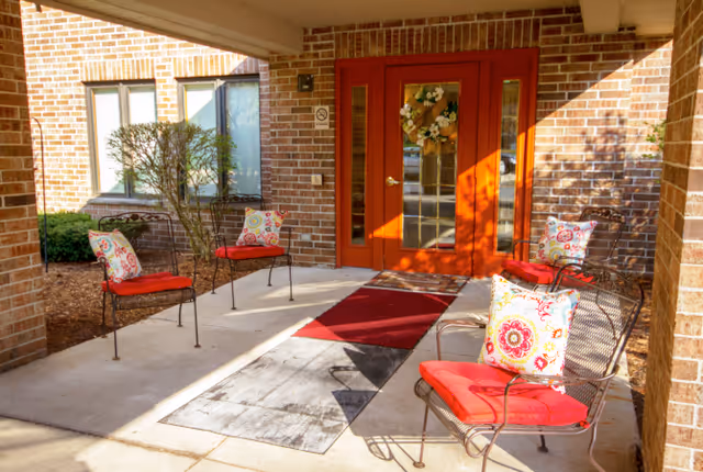 Covered brick entrance with red double doors and a porch furnished with metal chairs featuring red cushions and patterned pillows.
