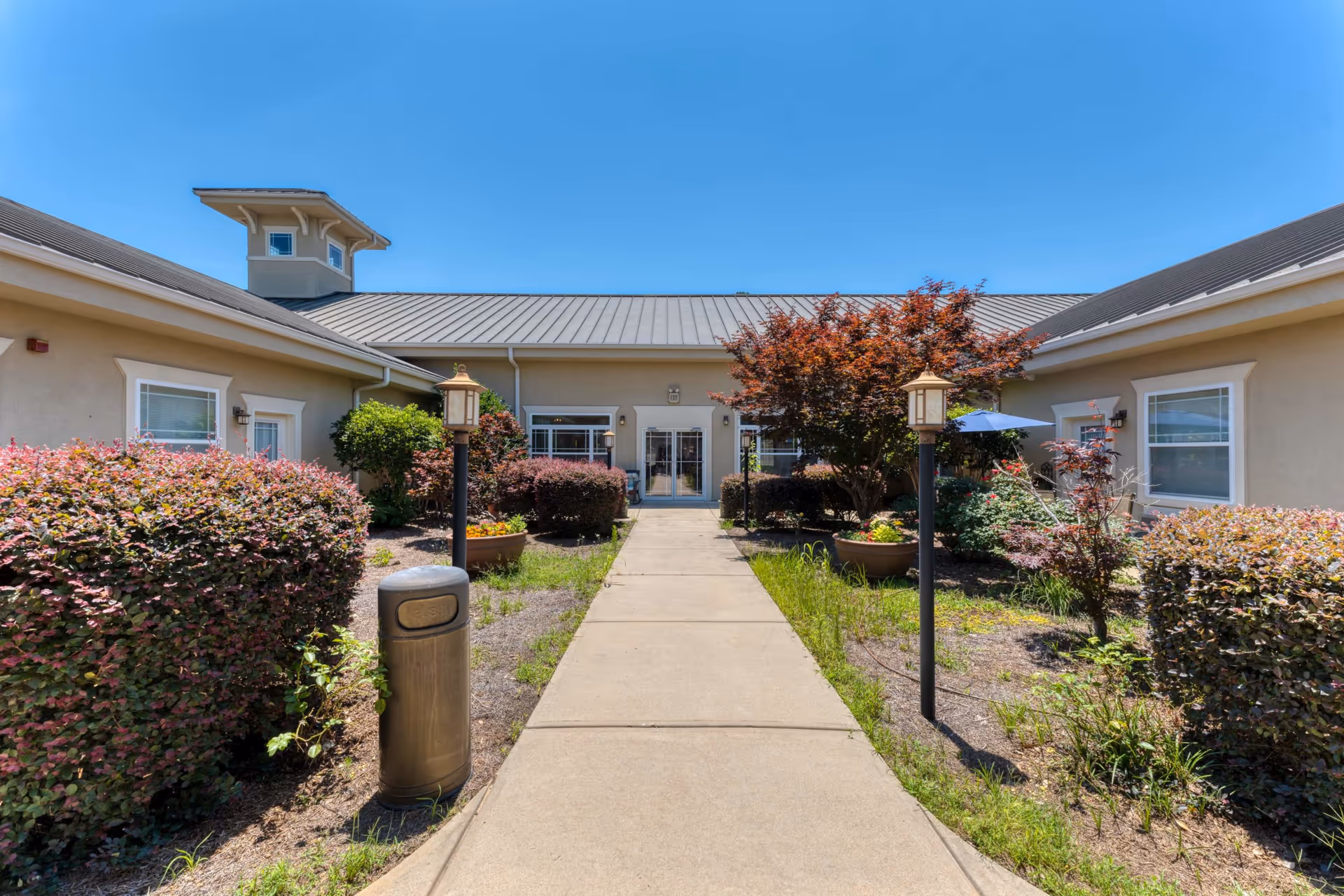 A paved walkway through a landscaped courtyard leading to the front entrance of a single-story senior living building under a clear blue sky.