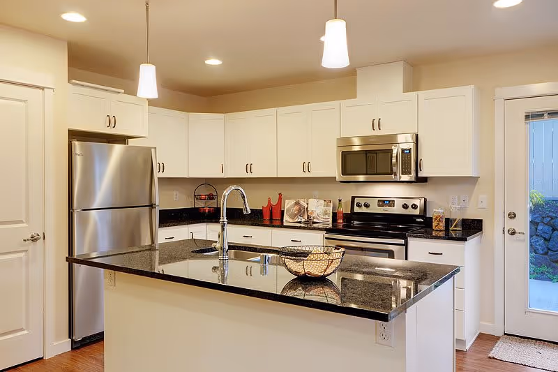 Modern kitchen with white cabinets, stainless steel refrigerator, oven, and microwave. A black granite countertop island with a sink and faucet is in the center, with pendant lights hanging above. A glass door leads outside, and the floor is wooden.