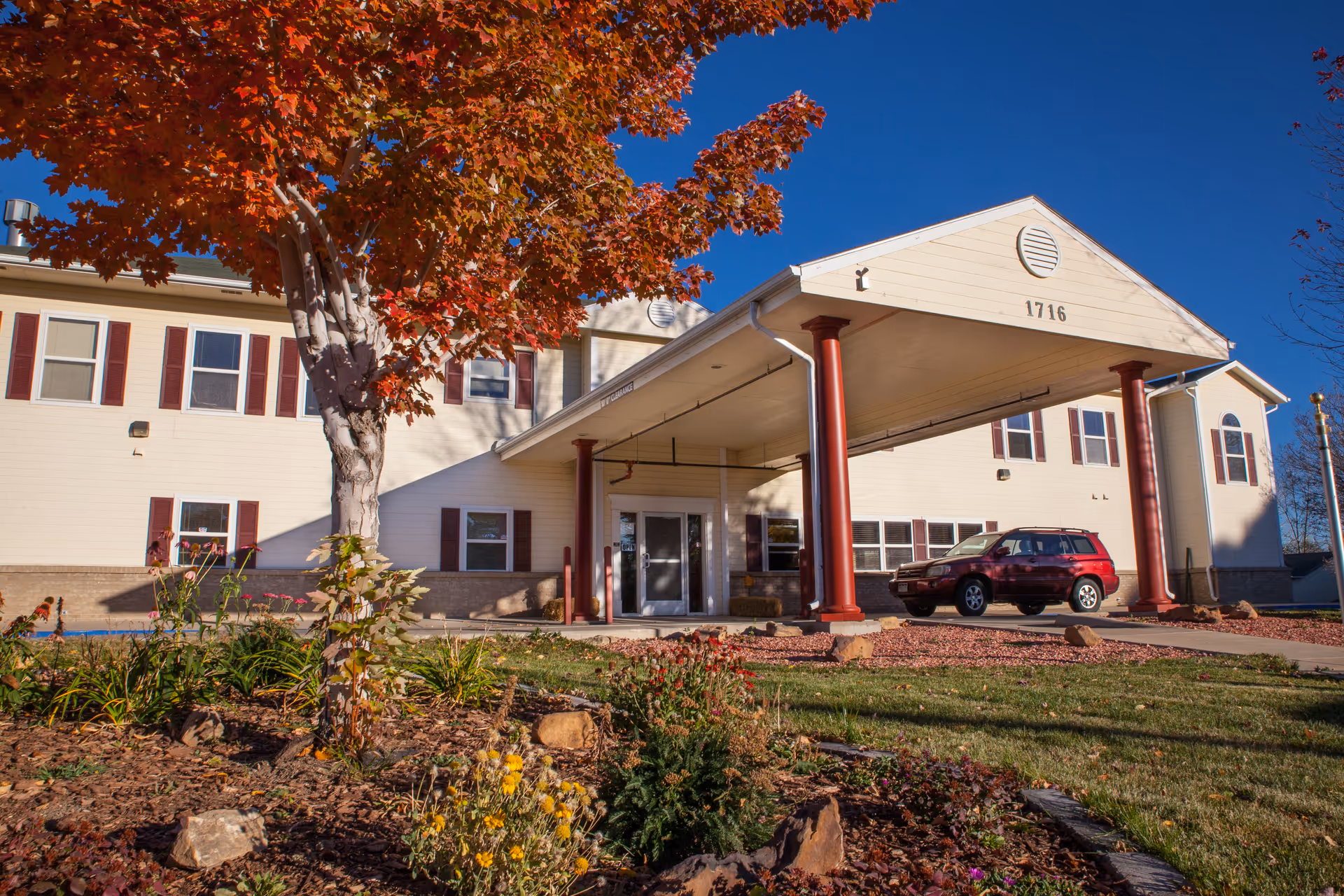 Front entrance of a two-story senior living building with a covered porte-cochere, red columns, an autumn tree, and a parked red SUV.