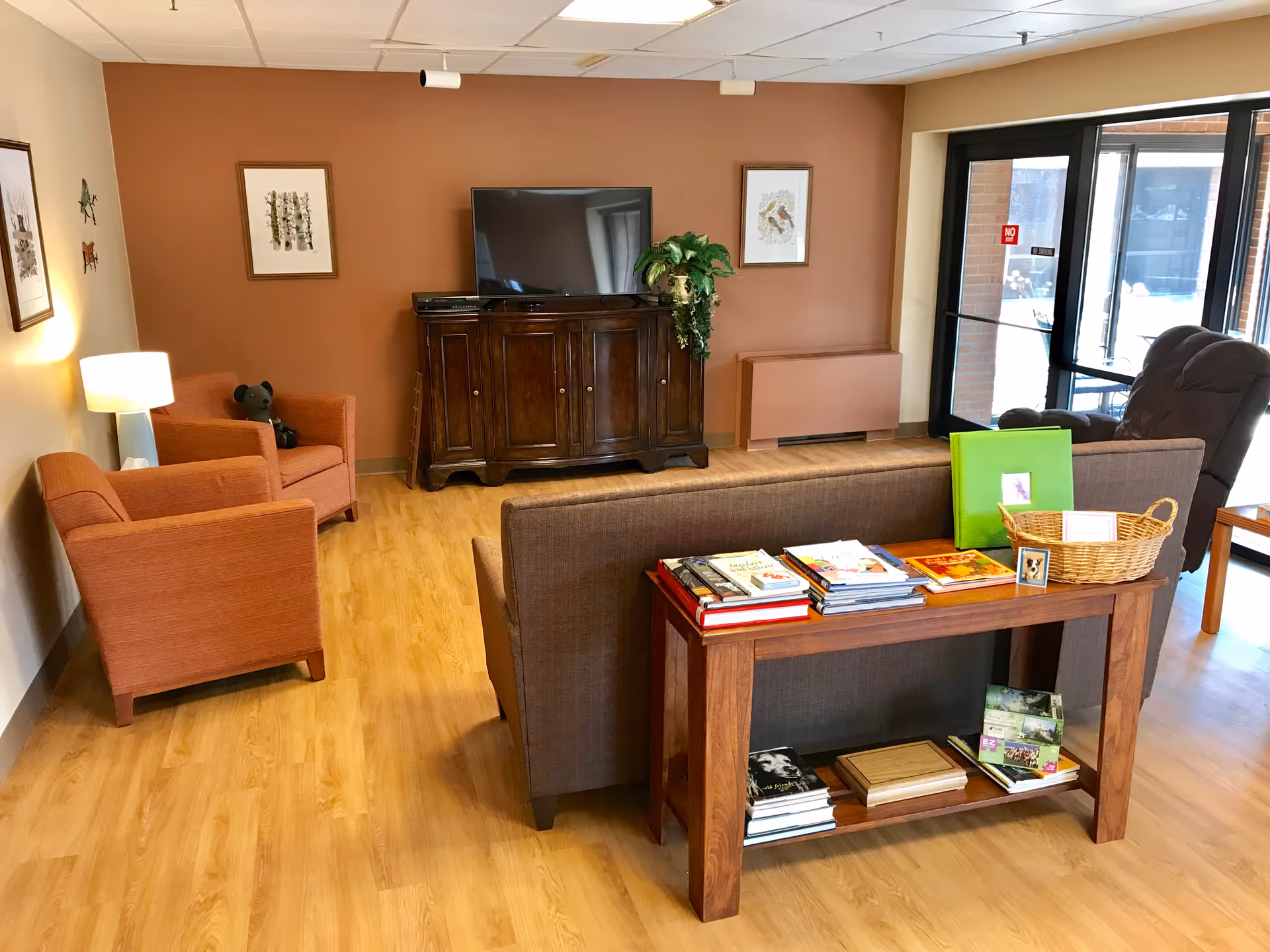 Cozy senior living lounge with sofa, armchairs, magazines, TV, and glass entry doors.