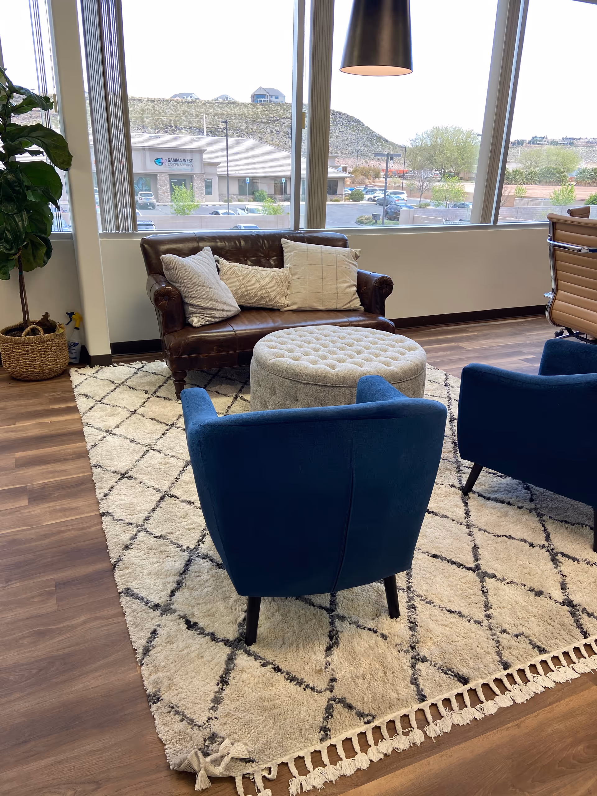 Cozy living room with blue chairs, a tufted ottoman, and a brown leather sofa by windows.