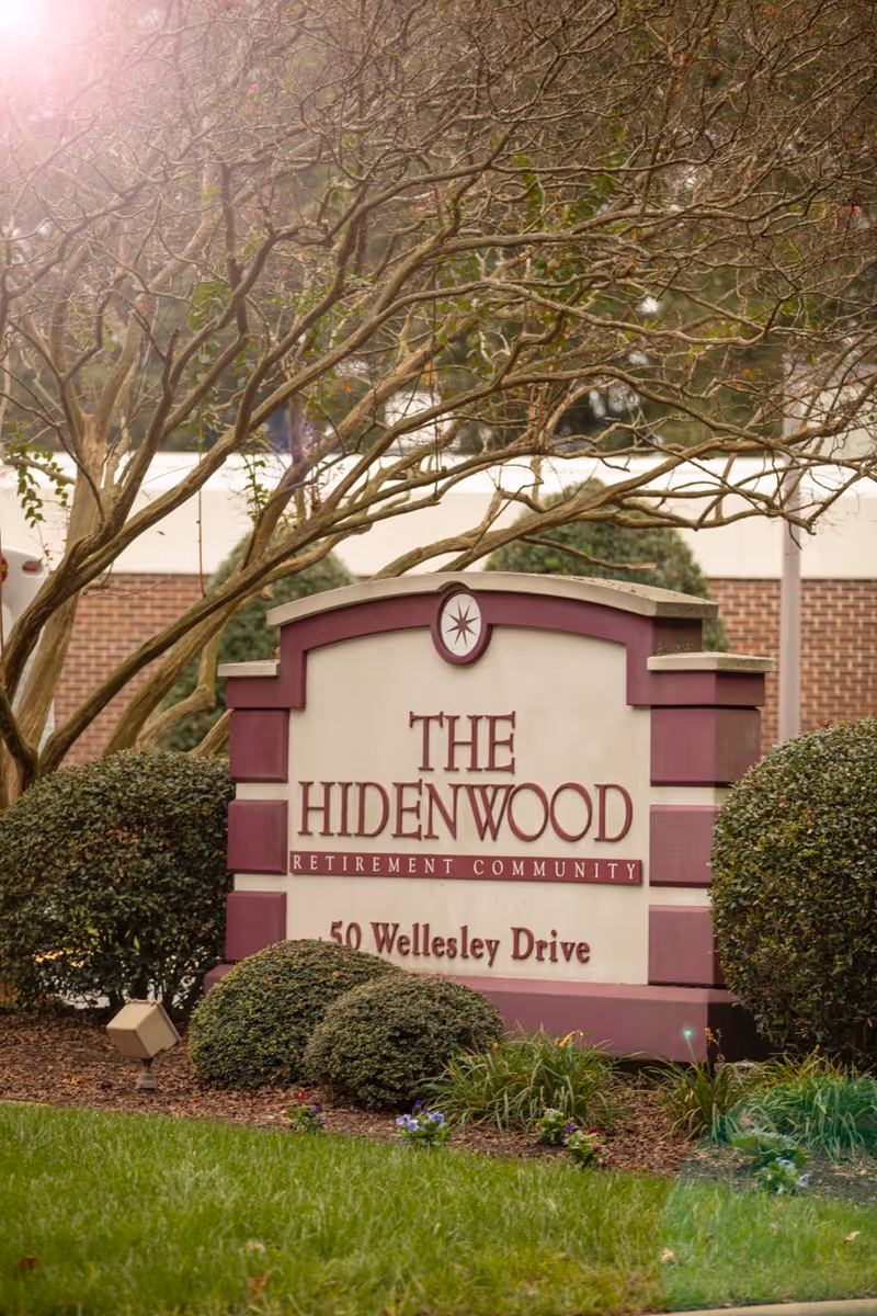 A large sign for The Hidenwood Retirement Community at 50 Wellesley Drive, surrounded by neatly trimmed bushes and a tree with bare branches, with a brick building partially visible in the background.