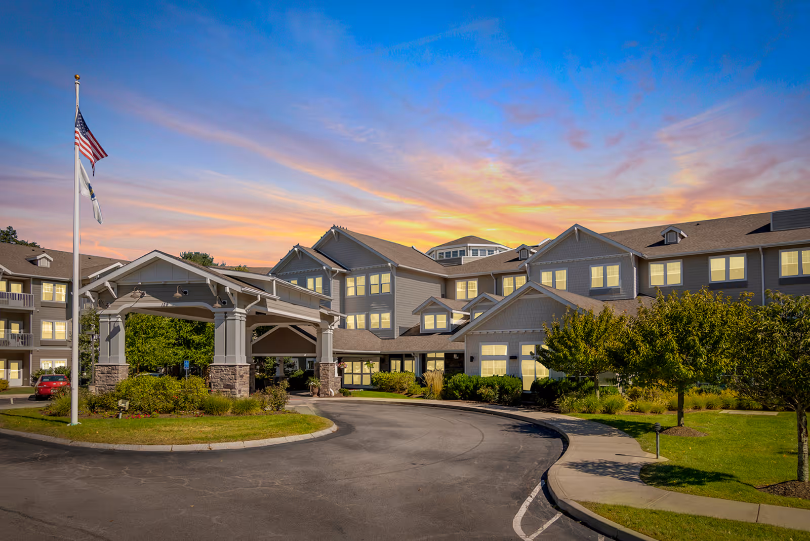 Exterior front of a multi-story senior living building with a covered entrance, flagpole, landscaped driveway, and a colorful sunset sky.
