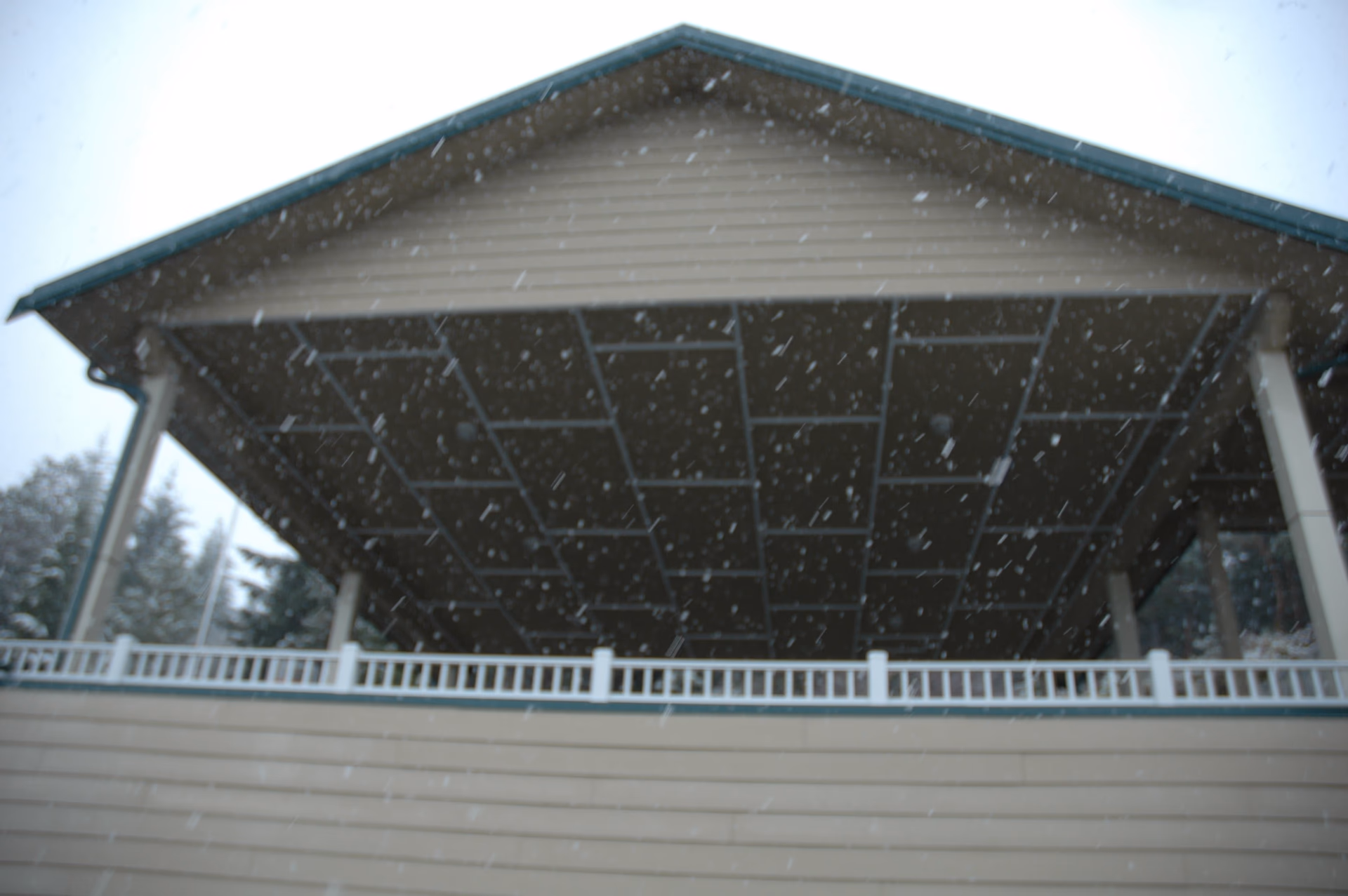 Snow falling beneath a large covered porch/overhang with white railing on the front of a building.