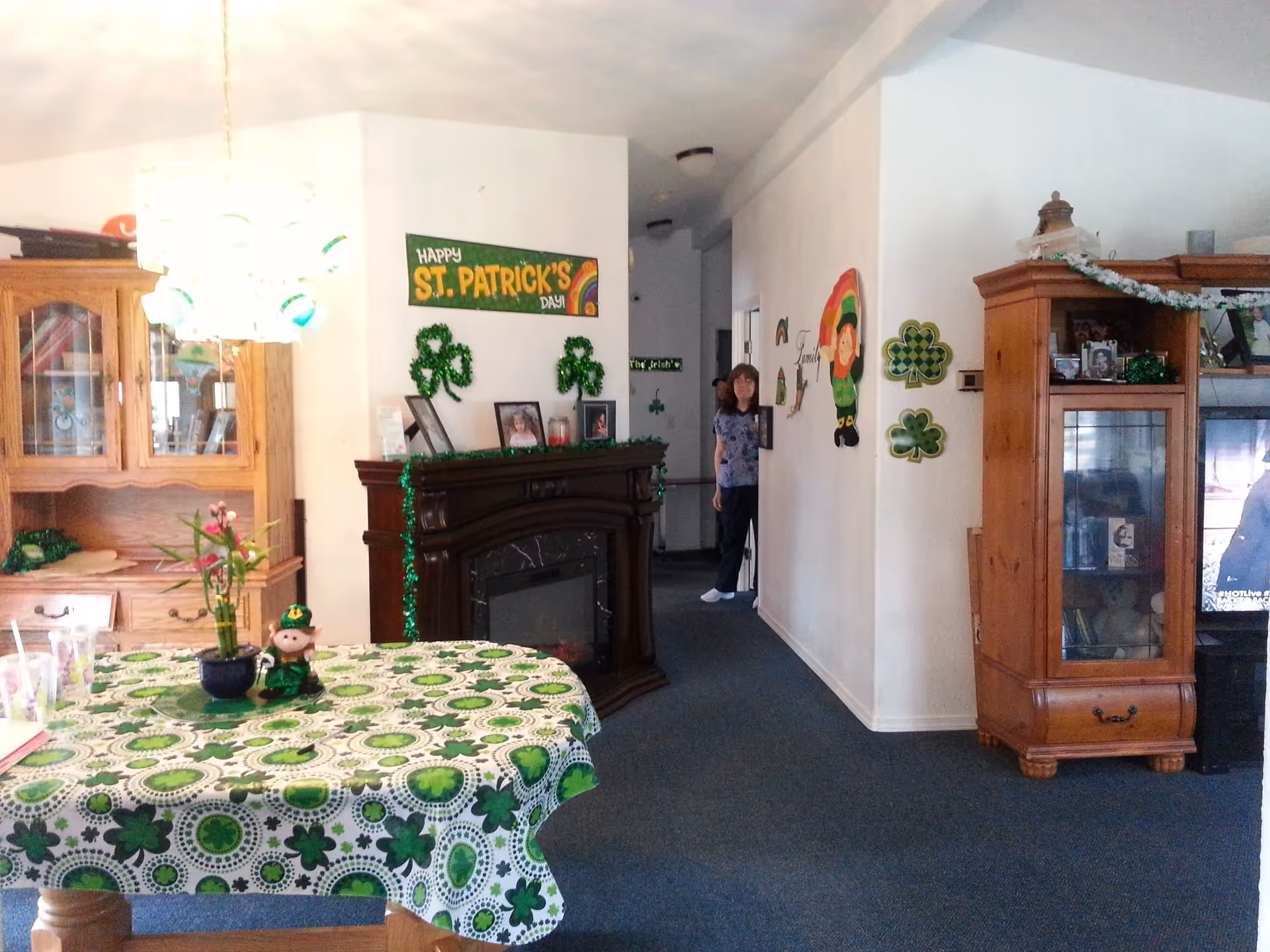 Interior view of a living room decorated for St. Patrick's Day with shamrock-themed tablecloth, wall decorations, and a 'Happy St. Patrick's Day' banner. There is a wooden cabinet, a fireplace with framed photos, and a person standing in the hallway.
