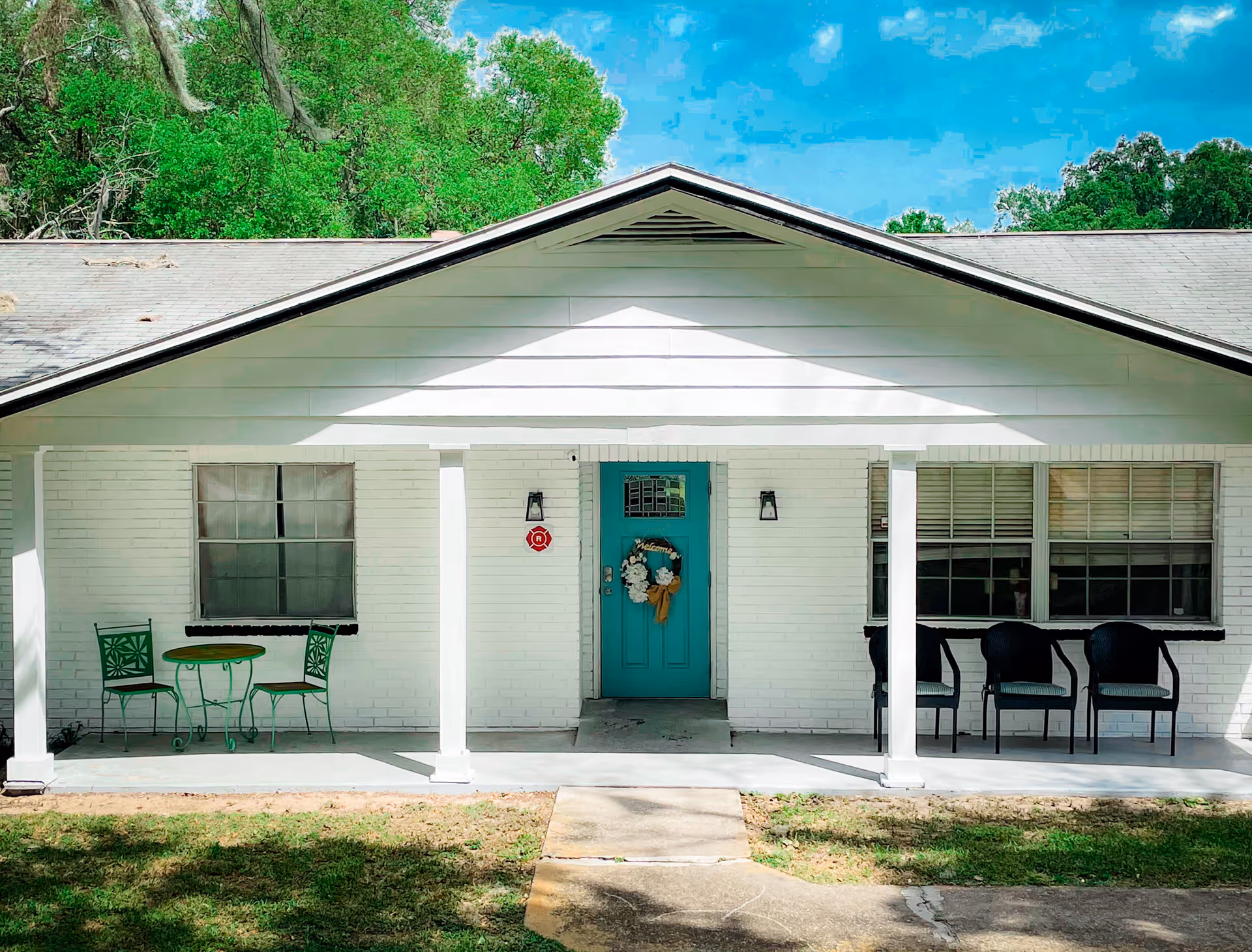 Front exterior view of a single-story white brick building with a teal front door decorated with a wreath. The porch has white columns, two black wall lanterns, a small green table with two matching chairs on the left, and three black chairs on the right. There are two windows on either side of the door, and green trees are visible in the background under a blue sky.