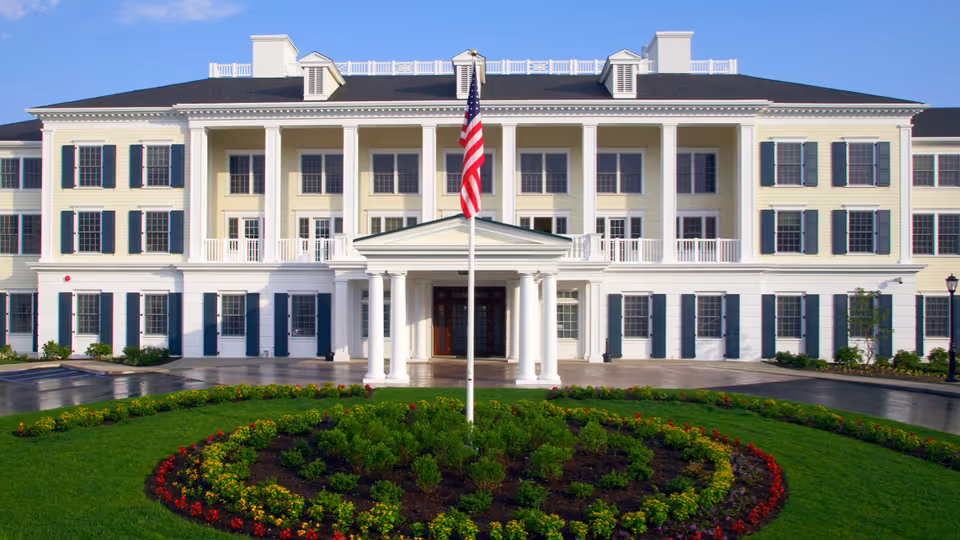 Large white, columned senior living building with an American flag and circular flower bed.